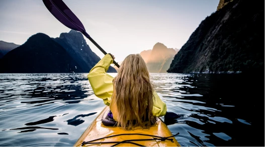 A woman in a kayak on the water in New Zealand, with beautiful mountains in the distance.